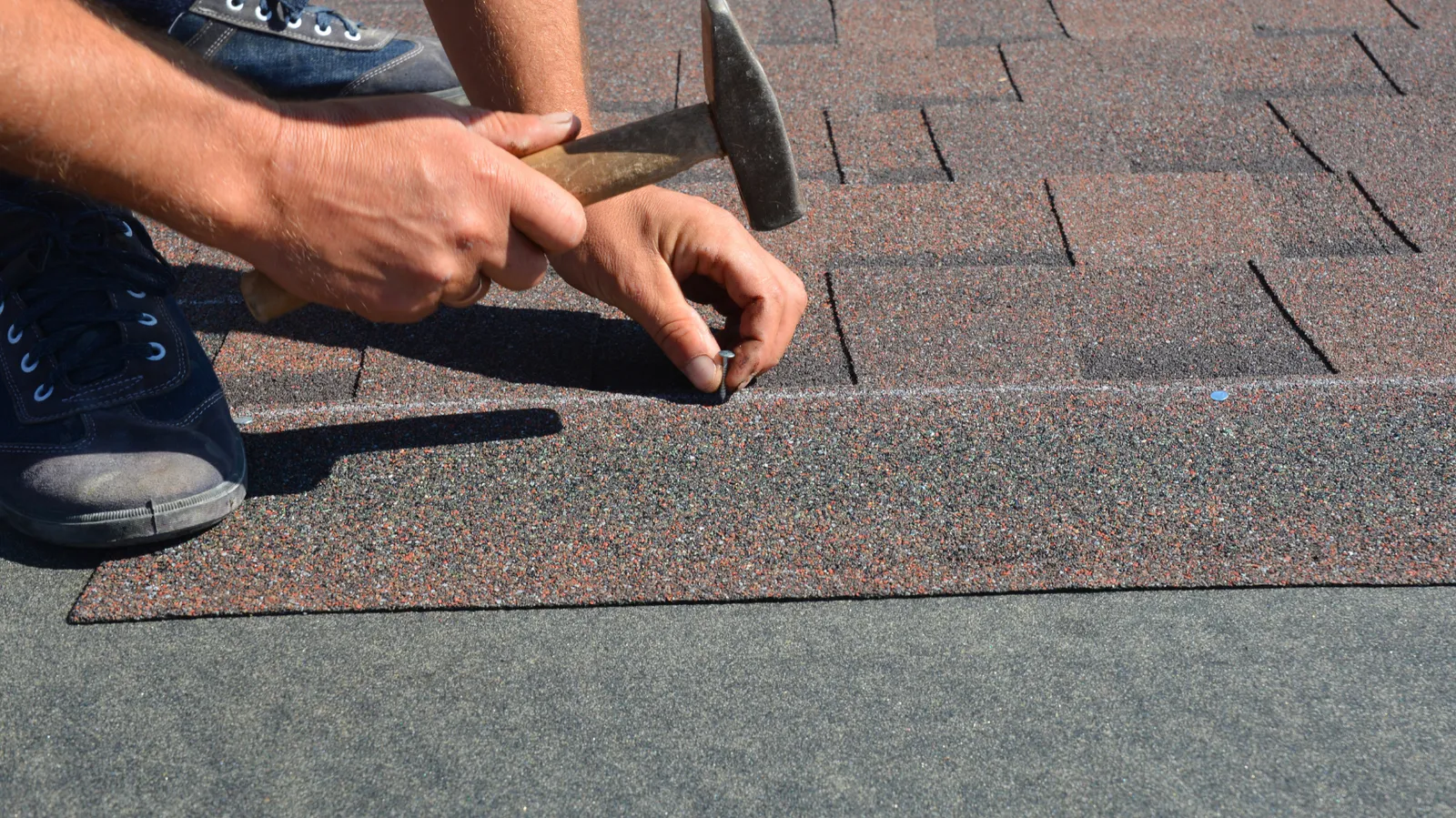 An asphalt shingle being nailed with a hammer by a roofer